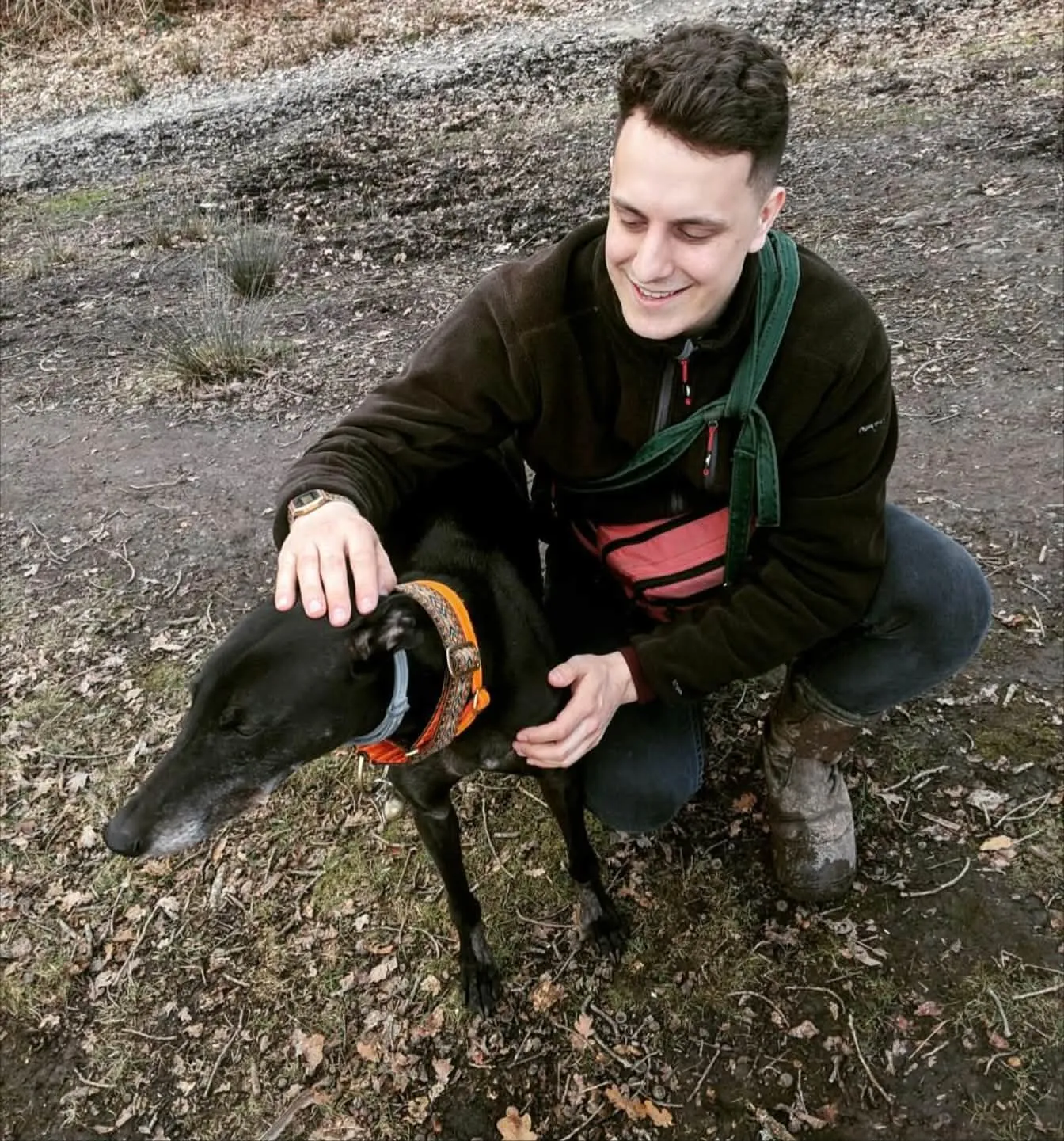 Joe crouching on a woodland path, stroking a black greyhound